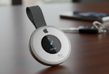 A conceptual second-generation Apple AirTag 2with a circular metal body, a small central digital display showing a tracking arrow, and an integrated fabric strap, resting on a wooden table